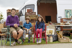 Man, boy and girl sitting on vintage chairs in front of a bric-a-brac stall with trunks and baskets and furniture on display at a flea market.